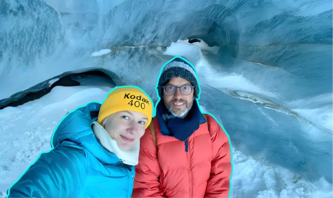 Selfie d’un couple en doudounes bleu et rouge, bonnet jaune, dans une grotte de glace bleutée.