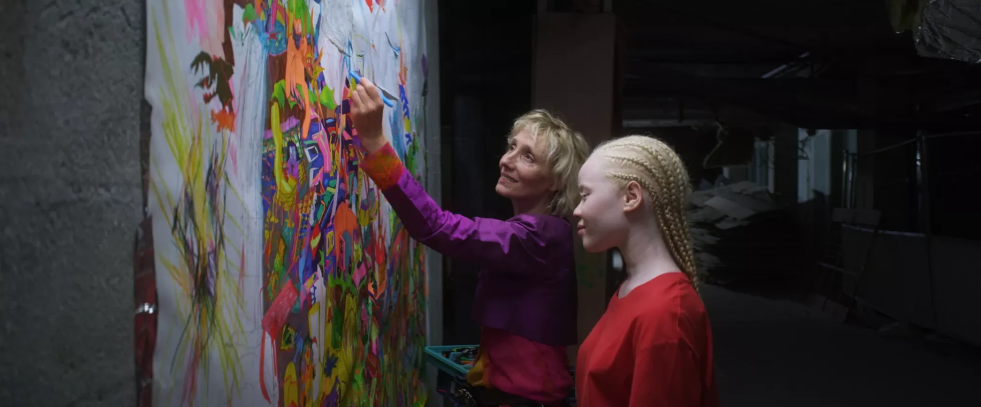 Femme en violet peint une fresque multicolore sur un mur, sous le regard d’une fille tressée en rouge, en intérieur.