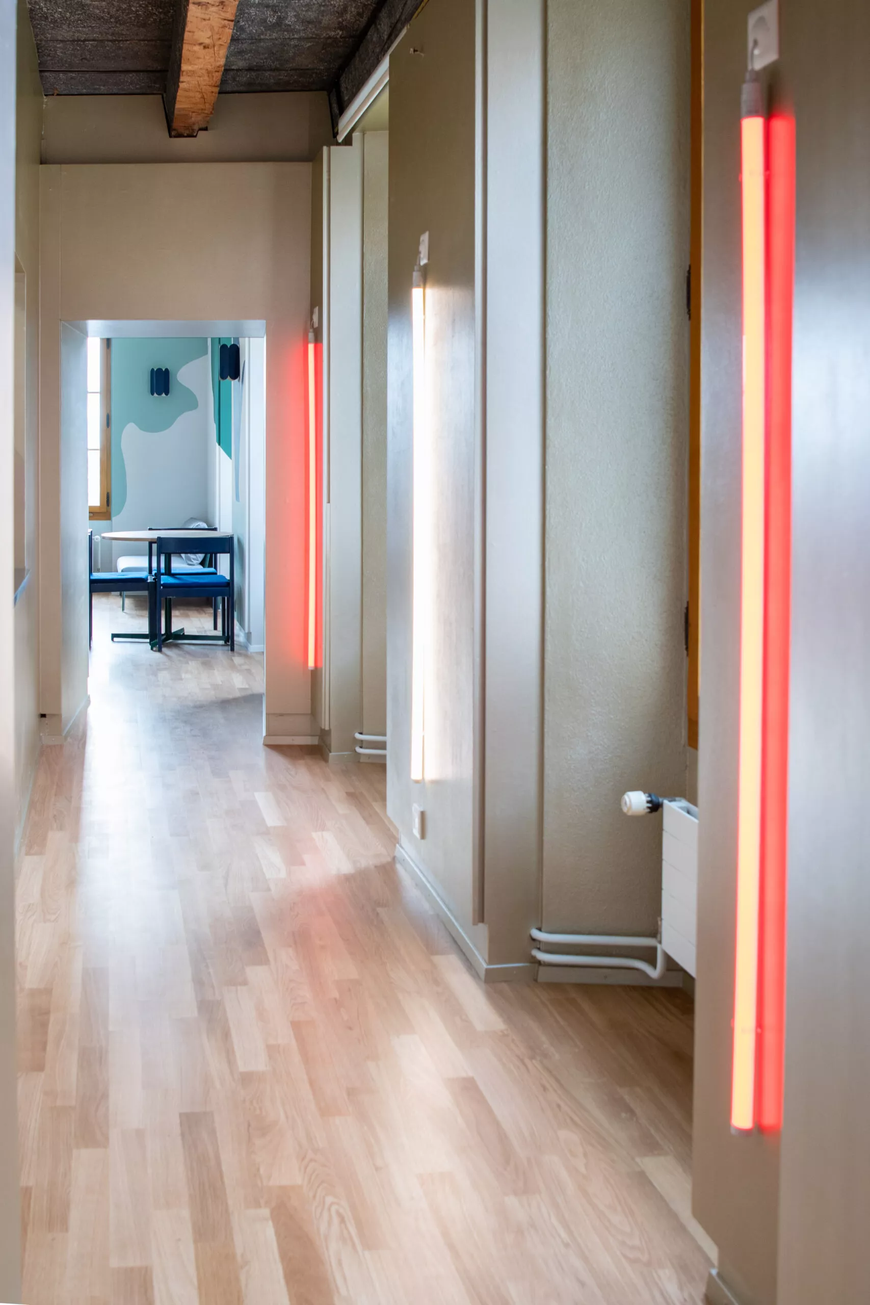 Couloir aux murs beiges, parquet clair, néons verticaux rouges; au fond, salle avec table, chaises bleues et fresque.