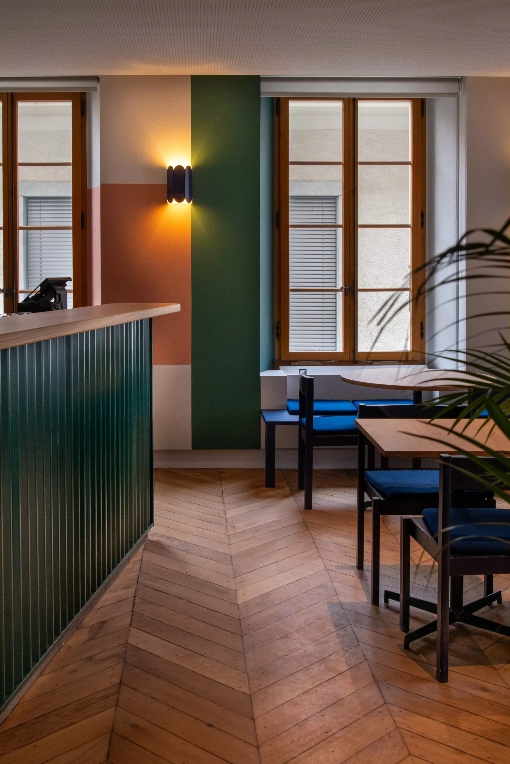 Intérieur du Pyxis Café : parquet en chevrons, mur vert et terracotta, appliques, tables bois et banquettes bleues.