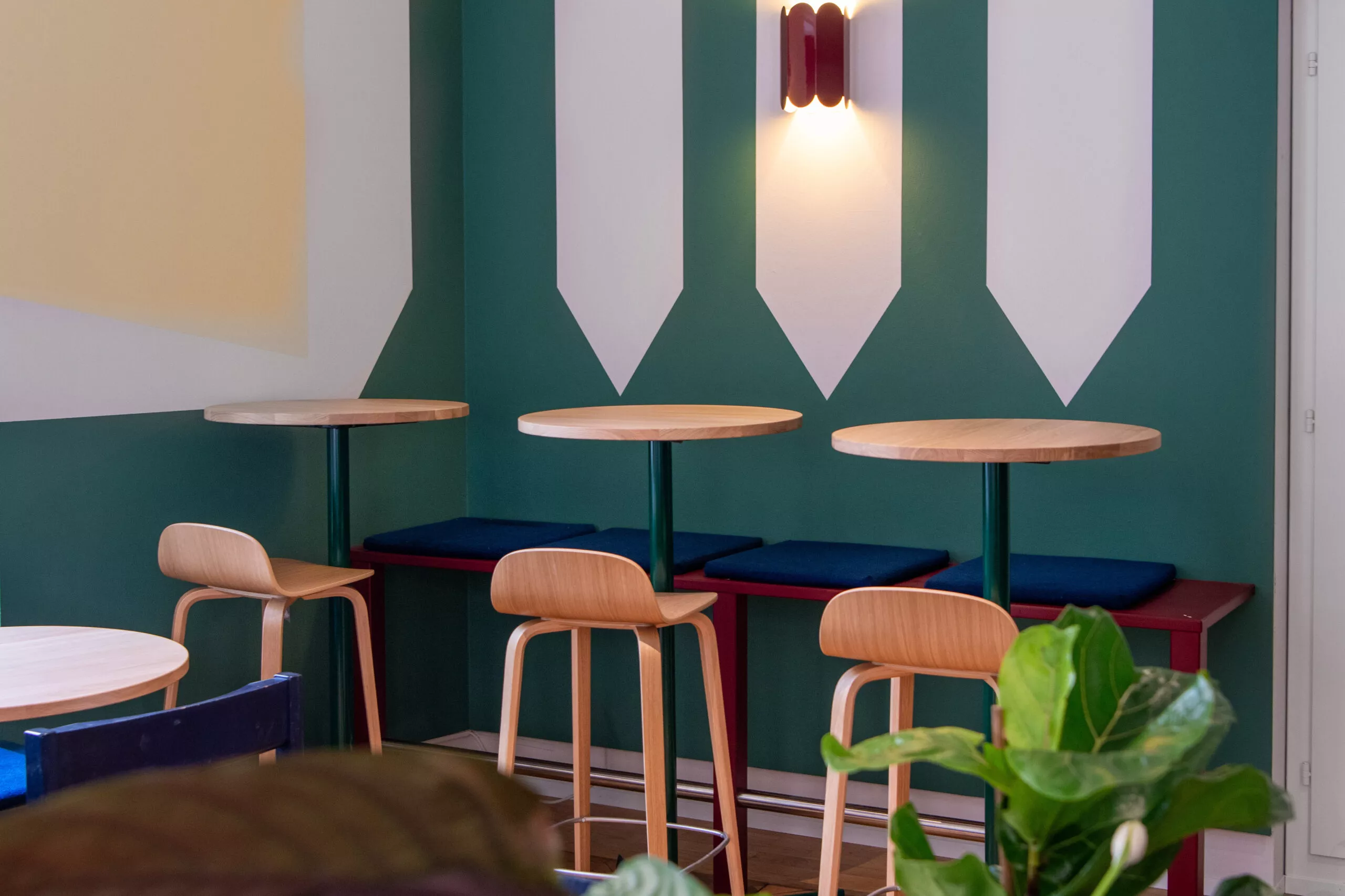 Coin café moderne: mur vert et blanc à motifs, tables rondes en bois, tabourets, banquette bleue, plante.