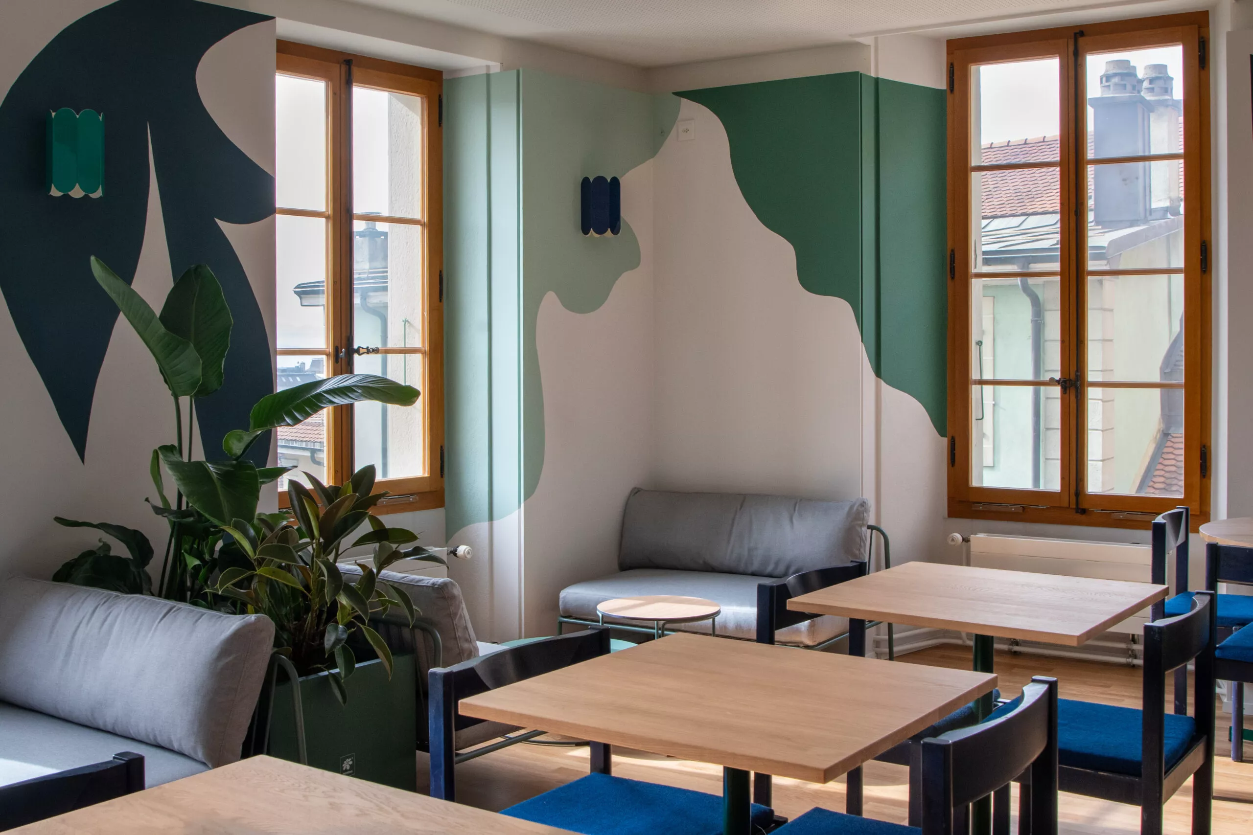 Salle du Pyxis Café lumineux: tables en bois, chaises bleues, canapés gris, plantes et mural vert/bleu, grandes fenêtres.